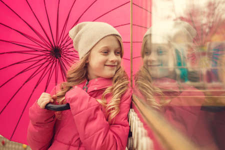 Cute little girl with umbrella looking on her reflection at window in parkの写真素材