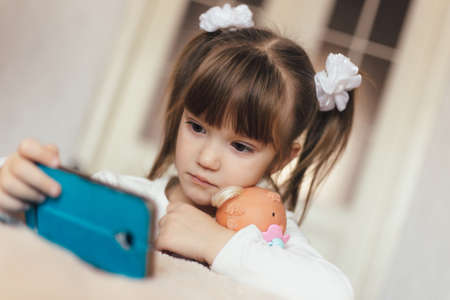 Little girl sitting near bed with mobile phone in living room at homeの写真素材