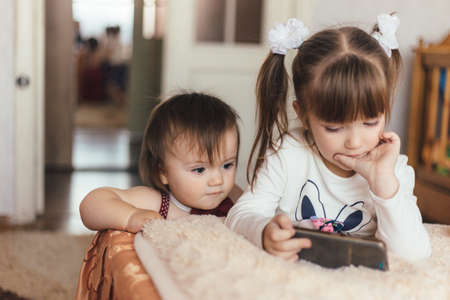 Little brother and sister standing near bed with mobile phone in living room at homeの写真素材