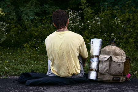 Back view of hiker with backpack sits on road next to forest