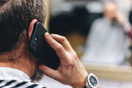 Back view of man talking on the smart phone and sitting on a chair in front of mirror at the barber shop. Shallow depth of field.の写真素材