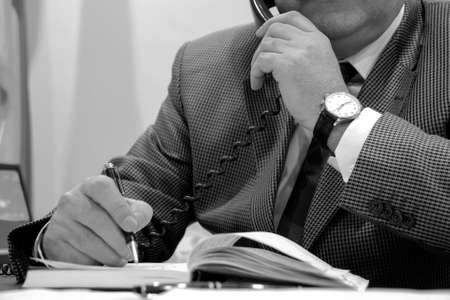 Businessman sitting at his desk making notes and talking on telephone. Shallow depth of field.の写真素材