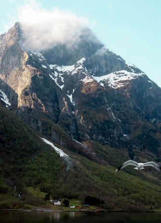 Giant rock with snow and small village on bank of fjord, Norway. Sunny day, blue sky with white...