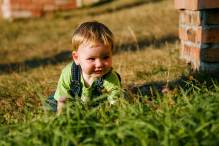 Lutsk, Volyn / Ukraine - September 27 2011: Boy little child with thoughtful face with blond hair in blue jeans crawling on his knee on park with green grass in summer day. Shallow depth of field.のeditorial素材