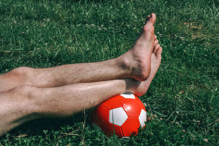 Barefoot legs on soccer ball. Man lying on grass of field. Outdoor at daytime on summer day. Healthy lifestyle concept.の写真素材