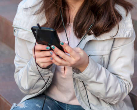 Young woman listening to music with earphones using smartphone sitting on stone staircase on street....