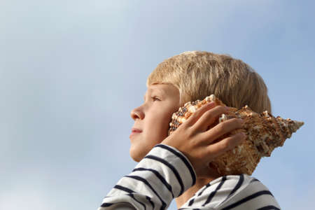 Happy caucasian boy listen to seashell at the sky background. Shallow depth of field.の写真素材
