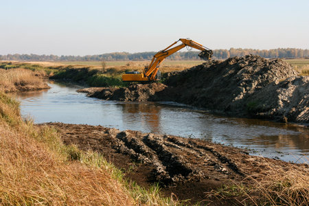 Heavy yellow excavator working on deepening and cleaning a small river, water running out from bucketの写真素材