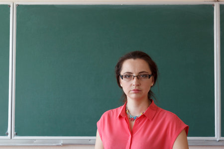Woman teacher with glasses stands in front of green school chalkboard in classroomの写真素材