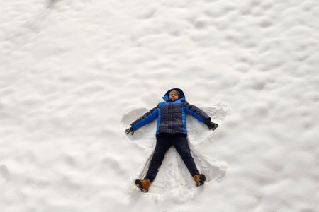 Teenager boy in blue jacket lying on snow and making angel