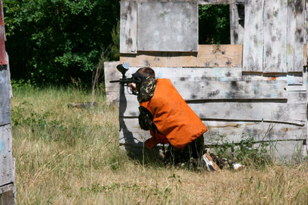 Rear view of male player in goggle mask and camouflage with paintball gun on a special playing field with fortificationsの写真素材
