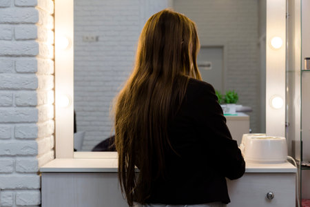 Back view of young woman with long hair posing in front of the mirror in beauty room. Shallow depth of field.の写真素材