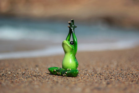 Green frog figure in yoga pose on sand of beach at sunny day. Shallow depth of field.