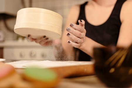 Woman sifts white flour through a wooden sieve on table at kitchen. Shallow depth of field. Cropped...