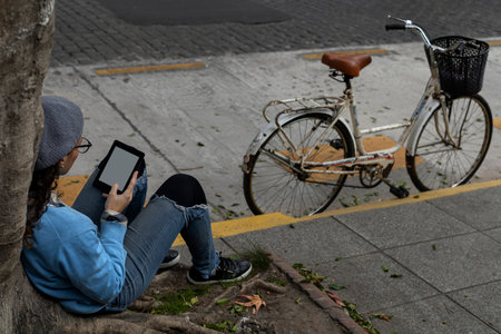 Young Latin American female hipster sitting on the sidewalk reading a digital bookの写真素材