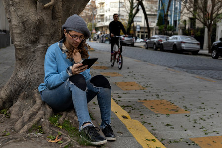 Young Latin American female hipster sitting on the sidewalk reading a digital book