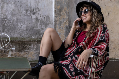 Young Latin American woman with curly hair, hat, sunglasses, positive attitude talks on cell phoneの写真素材