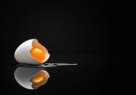 Broken eggshell with raw egg on a black minimalist surface reflective backgroundの素材