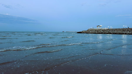 Calm blue seascape with lighthouse, low tide revealing rippled sand and shallow pools, jetty stretching toward beacon, overcast sky casting cool tones, distant gulls circling, scene evokes lighthouse