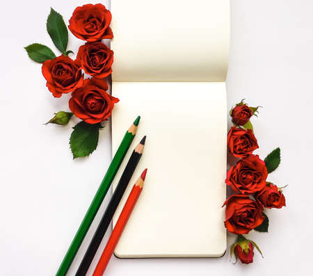 Colorful composition with sketchbook, red roses and pencils. Flat lay on white table, top view, copy spaceの写真素材