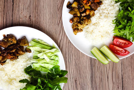 Top view of two plates with healthy homemade lunch. White rice, thai fried meat in sauce and colorful vegetables: rocket salad, cucumber, tomato, cabbage. FLat lay on wooden backgroundの写真素材