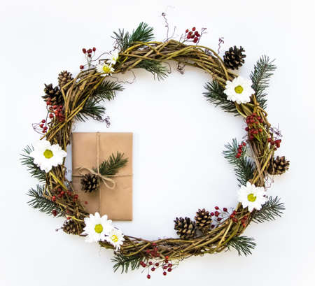 Festive wreath of grape vines, decorated with berries, fir branches, daisy flowers and cones. Present wrapped in eco paper. Christmas DIY wreath. New Year round wreath on white background. Flat lay, top viewの写真素材