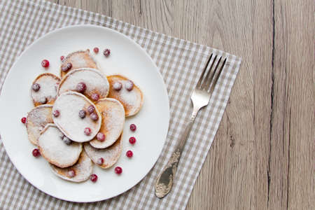 Home made pancakes with cranberries and sugar powder on a white plate. Wooden table, rustic decoration, berries and a fork. Top view, flat lay, view from aboveの写真素材