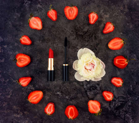 Colorful bright composition of make up accessories, strawberries and peony flower. Overhead view of lipstick and mascara. Flat lay, top view, cute feminine still life on dark stone backgroundの写真素材