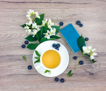 Spring bright composition of cup of green tea, notebook, blueberries and bouquet of jasmine flowers on wooden table. Rustic home composition. Flat lay, top viewの写真素材
