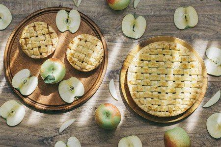 Dessert wooden plate with delicious homemade apple pies and set of whole fresh green apples and cut apple and slices on wooden texture background in soft natural light beaming. Top view.の写真素材
