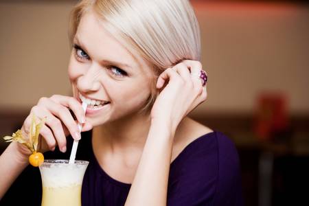 Playful blonde woman with a beautiful smile drinking a tropical cocktail through a straw, indoor portrait with copyspaceの写真素材