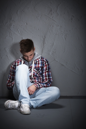 Young man feeling depressed sitting on the ground leaning against a wall and looking down wearing blue jeans and a checkered sweaterの写真素材