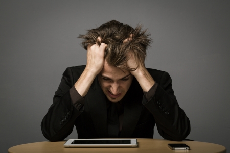 Man suffering from Burn Out sitting at his desk with his tablet-pc with his head bowed and hands running through his hairの写真素材