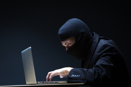 Computer hacker in a balaclava working in the darkness stealing data and personal identity information off a laptop computerの写真素材