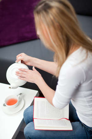 High angle view of a blond woman sitting reading a book stopping and pouring herself a cup of tea from a teapotの写真素材