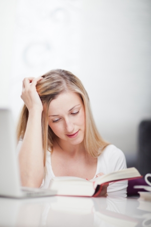 Young woman studying from a textbook sitting at a table concentrating on her reading with her head resting on her handの写真素材