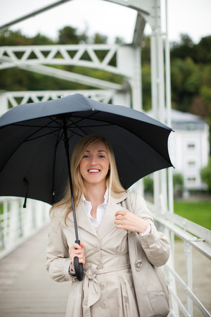 Woman out walking in the rain smiling as she shelters under her umbrella in her stylish beige raincoatの写真素材