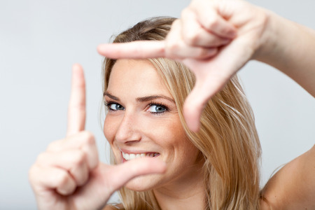 Playful vivacious young blond woman with a lovely smile framing her face with her fingersの写真素材