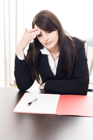 Businesswoman with a migraine headache sitting at her desk with on open folder of notes holding her hand to her forehead with a serious expression and downcast eyesの写真素材
