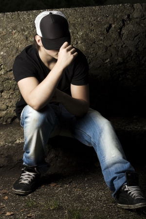 Depressed teenage sitting thinking in the late evening light with his baseball cap pulled low over his eyesの写真素材