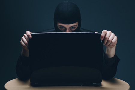 Furtive masked hacker accessing a laptop computer to steal data, plant malware or spy conceptual of...