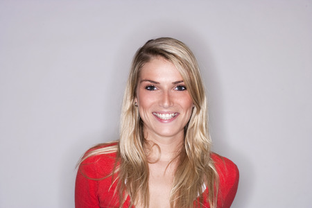 Beautiful blond woman with a radiant smile in a colourful red top posing on a grey studio background with copyspaceの写真素材