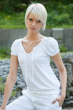 Beautiful slender young blond woman standing on a rock in a fresh white summer blouse and slacks as she enjoys a day out in the mountainsの写真素材