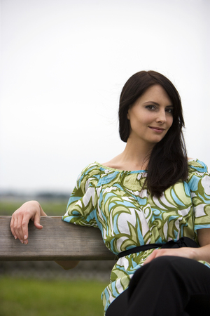 Beautiful stylish woman with a seductive smile relaxing outdoors on an old rustic wooden bench in a colorful blue and green top echoing the nature around herの写真素材