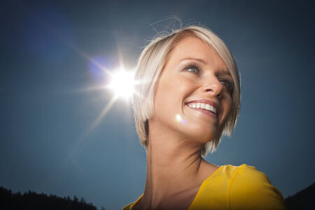 Kiss The Sun - a beautiful vivacious young blond woman has her cheek kissed by a bright sunburst as she laughs and smiles outdoors on a hot summer day against a clear blue sky, low angle viewの写真素材