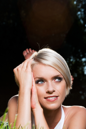 Beautiful tanned young woman lying on her stomach on green grass looking up into the sky with a dreamy look against a dark background with copyspaceの写真素材
