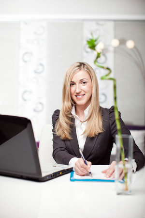 Beautiful businesswoman in a modern office sitting at her desk in front of a laptop computer writing notes on a pad and looking at the camera with a smileの写真素材