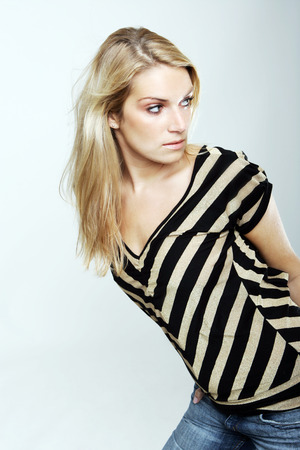 Attractive young woman with long blond hair in a striped black and white top standing leaning forwards looking behind her over her shoulderの写真素材