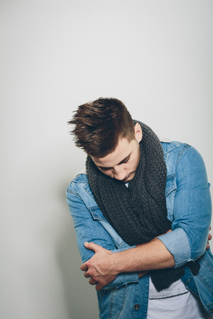 Sad Handsome Man Wearing Denim Jacket and Gray Knitted Scarf, Crossing his Arms in Front his Body While Looking Down. Isolated on Light Gray Background.の写真素材