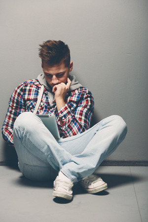 Contemporary casual young man in jeans and sneakers sitting cross legged on the floor using a tablet reading or studying information with a serious engrossed expressionの写真素材